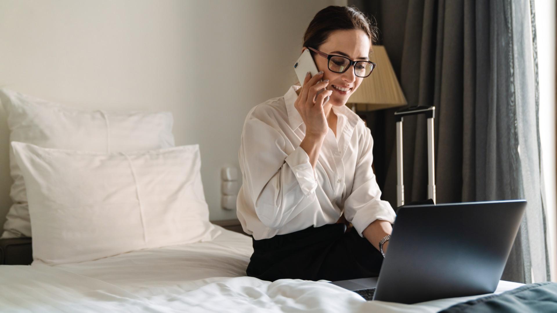 Donna al telefono sul letto con laptop, indossa occhiali e camicia bianca.