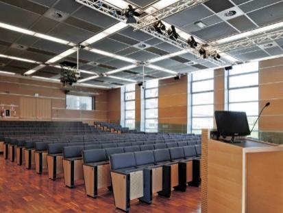 Aula conferenze moderna con sedili in legno, podio e illuminazione a soffitto.