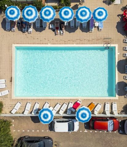 Piscina con lettini e ombrelloni blu, vista dall'alto, auto parcheggiate accanto.