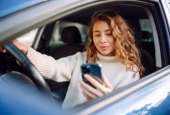 Donna in auto guarda il telefono, indossa un maglione bianco, guida distratta.