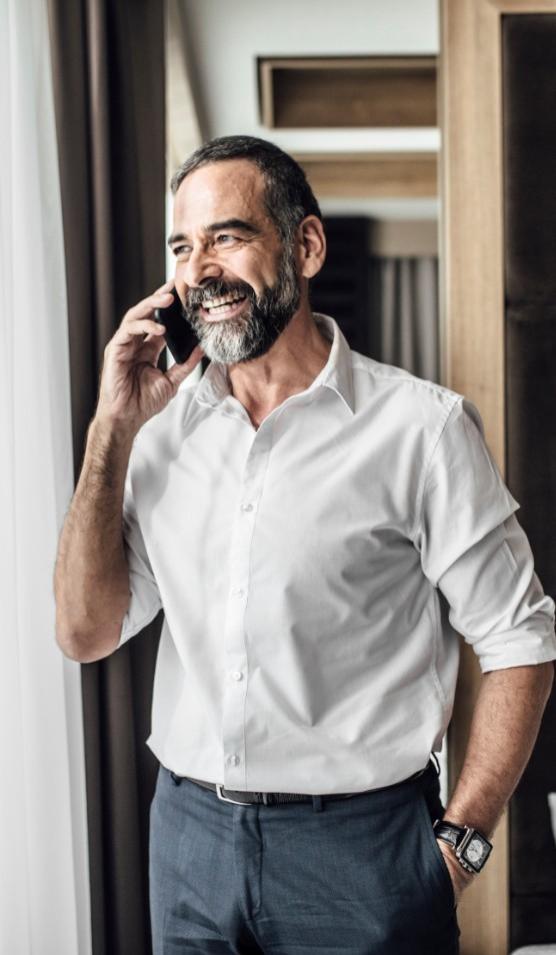 Uomo sorridente in camicia bianca parla al telefono vicino a una finestra.