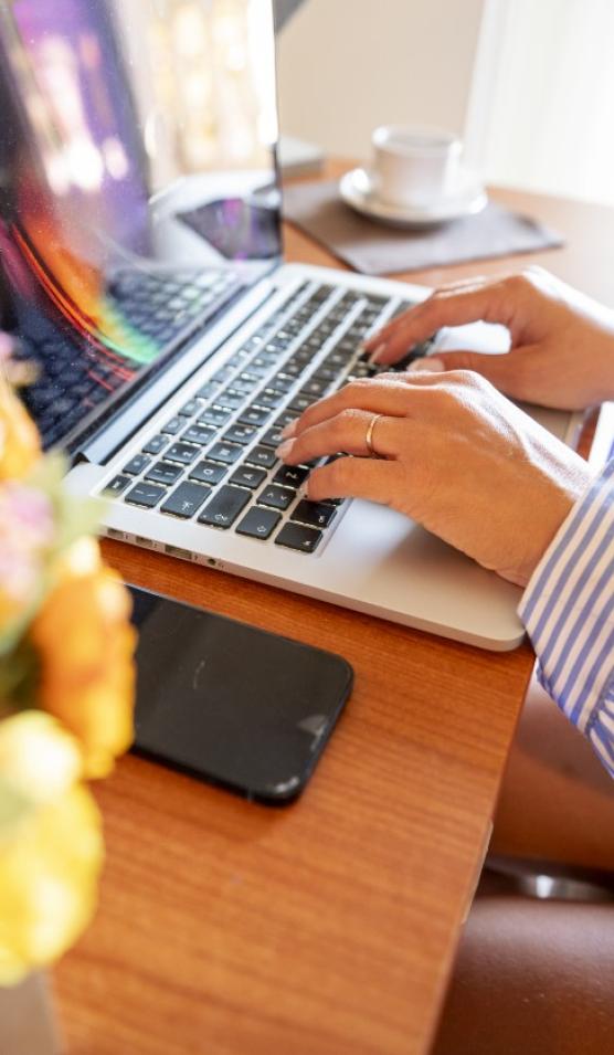 Persona al computer, smartphone vicino, tazza di caffè e fiori sul tavolo.