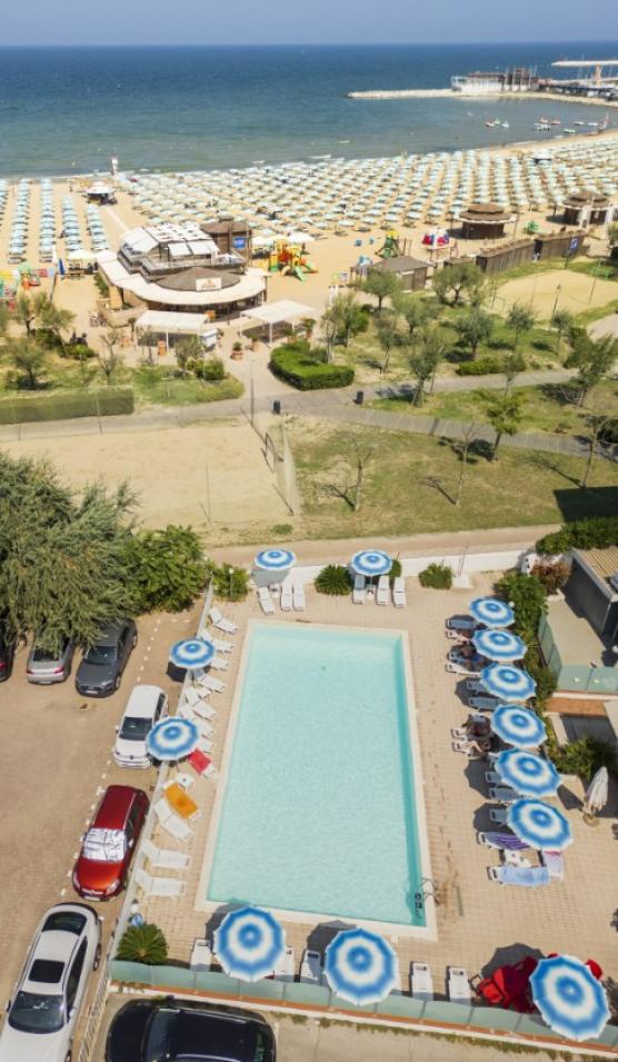 Spiaggia affollata, piscina con ombrelloni blu, parcheggio adiacente, vista mare.