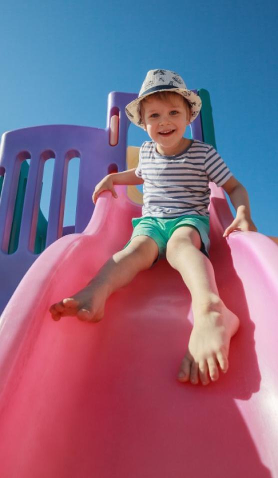 Bambino felice scivola su uno scivolo rosa sotto il cielo blu.