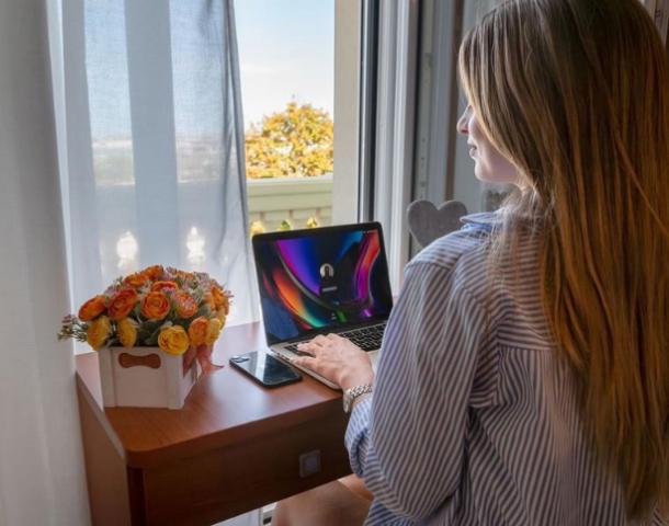 Donna al computer vicino a una finestra, con fiori e smartphone sul tavolo.