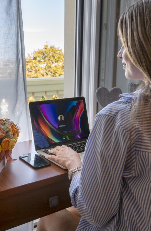 Donna lavora al laptop vicino alla finestra con vista esterna e smartphone sul tavolo.