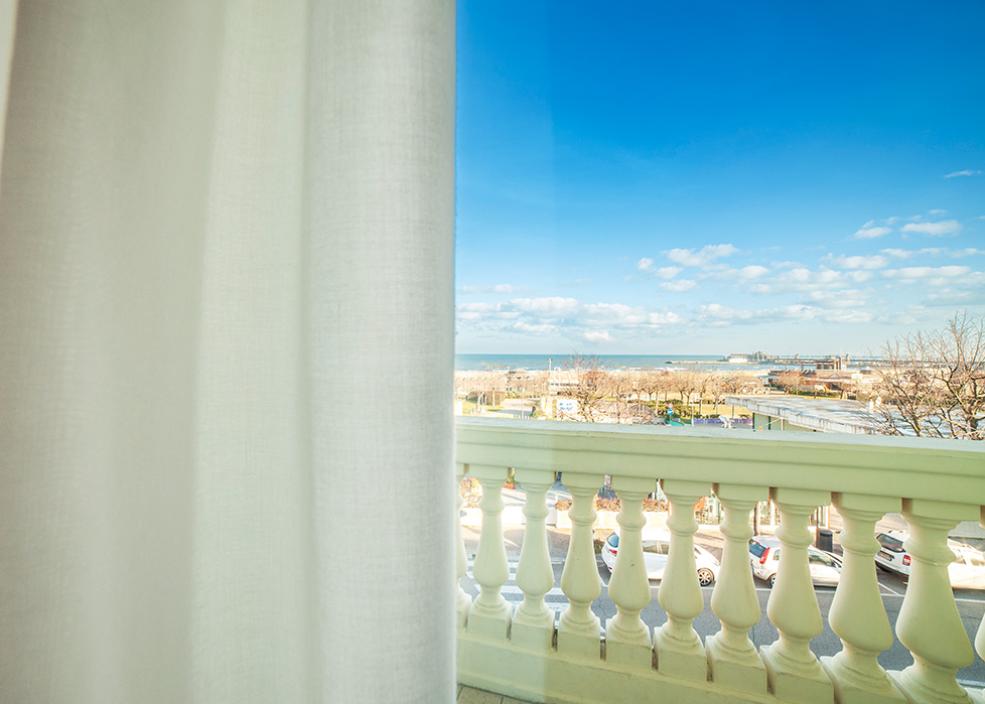 Vista dal balcone con mare e cielo blu, parzialmente coperta da una tenda bianca.