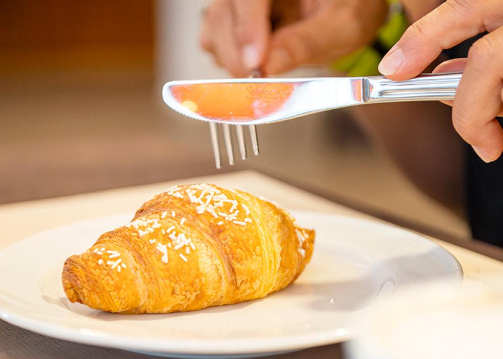 Croissant su piatto bianco, persona con forchetta e coltello, tazza di caffè accanto.