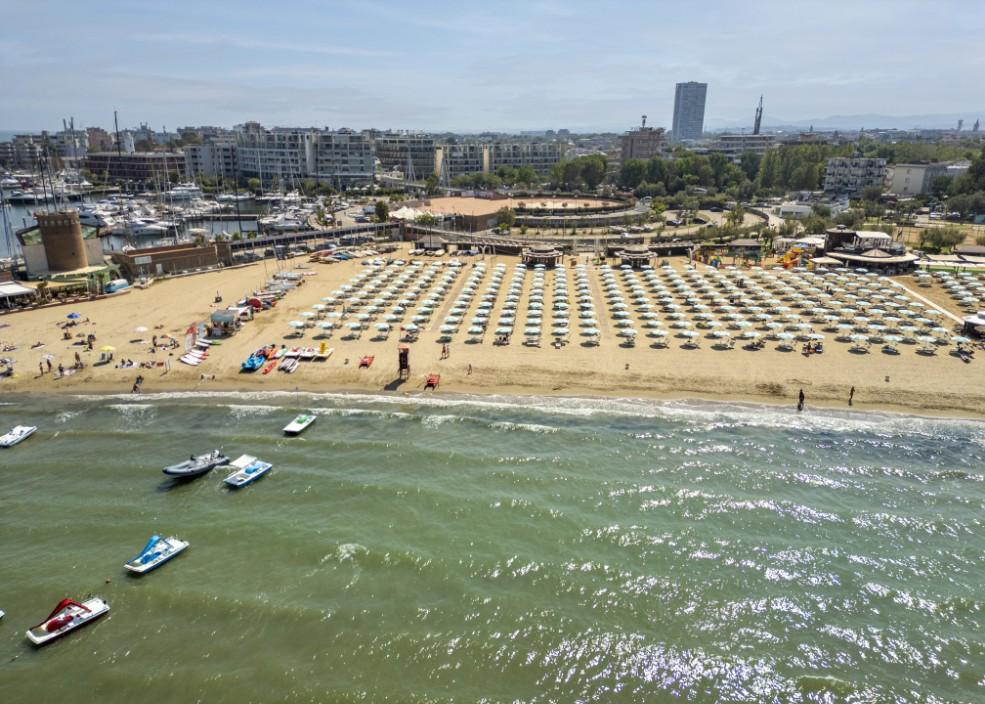 Spiaggia affollata con ombrelloni, barche e vista sulla città sullo sfondo.